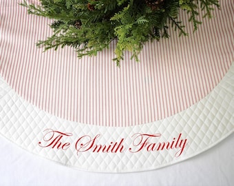 May include: A round Christmas tree skirt with a cream and red striped pattern. The border is quilted white fabric. The words "The Smith Family" are printed in red script. Greenery and pinecones are at the top.