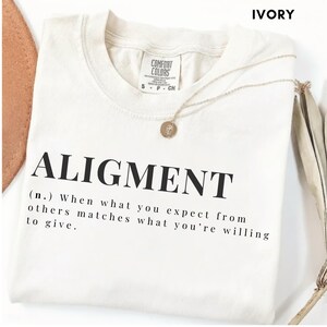 May include: An ivory short-sleeved t-shirt featuring the black printed text ALIGMENT followed by the definition: (n.) When what you expect from others matches what you're willing to give. The shirt has a Comfort Colors brand label at the neck.