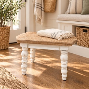 Peut inclure: Un petit tabouret en bois avec un plateau texturé brun clair naturel et quatre pieds tournés peints en blanc. Un textile tissé de couleur crème plié, avec de fines rayures brun foncé et des bords à franges, repose sur la surface du tabouret.