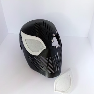 May include: A black full-face mask featuring a geometric lattice pattern across the forehead and mouth area. One white mesh eye lens is attached to the mask, while a second matching white mesh eye lens is positioned separately on the surface.