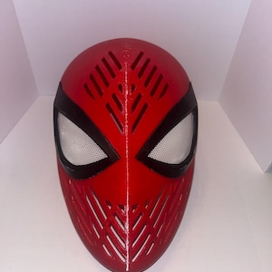 May include: A red full-face mask featuring a series of diagonal ventilation slits across the forehead and chin areas. The mask includes two large, almond-shaped eye openings outlined in black, filled with a fine white mesh material.