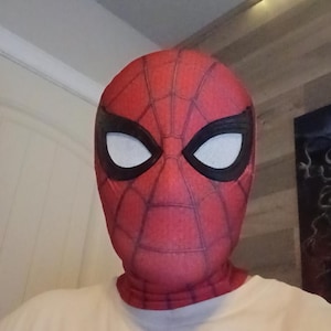 Peut inclure: Un masque en tissu rouge avec un motif de toile d'araignée noire et de grands verres oculaires blancs bordés de noir. Le masque couvre la tête et le cou, porté avec un t-shirt crème affichant le texte Marvel dans un cadre noir et Spider-Man en rouge.