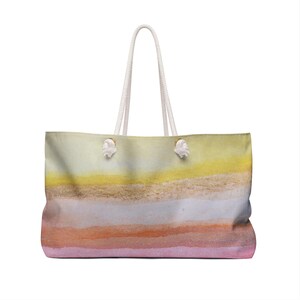 May include: A large rectangular tote bag featuring a horizontal watercolor gradient pattern. The design transitions from a pale yellow at the top, through shades of beige, white, and soft orange, to a light pink at the base. It has two white rope handles.