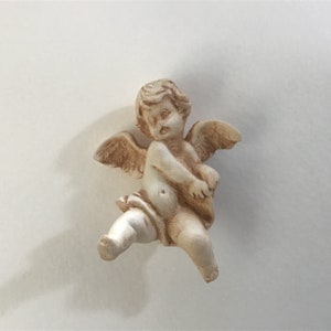 May include: A white ceramic cherub figurine with wings, holding a small object in its hands. The cherub has a sweet expression on its face.