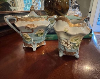 May include: A porcelain sugar bowl with a lid and a matching creamer pitcher, both featuring a light blue glaze, hand-painted white lotus flower motifs, and ornate gold trim along the scalloped rims, handles, and four small feet.