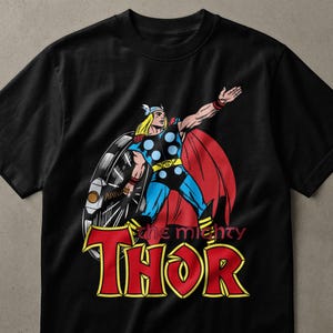 May include: A black short-sleeved t-shirt featuring a graphic of the comic book character Thor with blonde hair, a winged helmet, blue and black armour, and a red cape. The text "the mighty THOR" is printed in large red and yellow letters below.