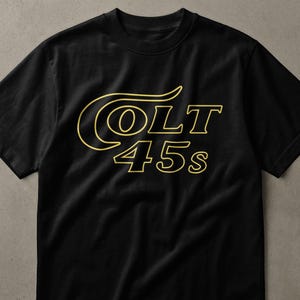 Puede incluir: Una camiseta negra de manga corta con el texto COLT 45s impreso en una fuente de contorno de color dorado en el pecho. El diseño incluye una C estilizada que se extiende sobre la palabra COLT, con el número 45s posicionado directamente debajo.