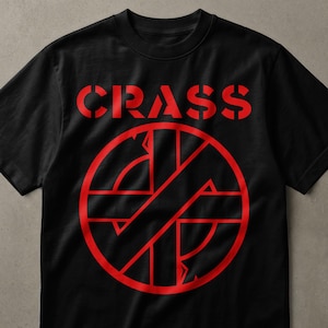 May include: A black short-sleeved t-shirt featuring the word CRASS printed in a bold red stencil font across the chest, positioned above a large red circular graphic containing a stylized cross symbol with intersecting lines and geometric shapes.
