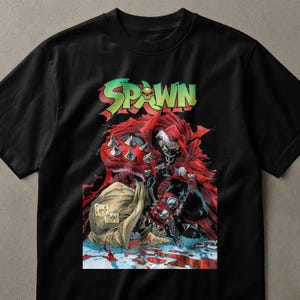 Könnte beinhalten: Ein schwarzes Kurzarm-T-Shirt mit einem Grafikdruck der Comicfigur Spawn. Das Design zeigt das Wort SPAWN in grünen Buchstaben mit einem Totenkopf-Symbol, einem roten Umhang, einer stacheligen Rüstung und einem Sack auf einem blutigen Boden.
