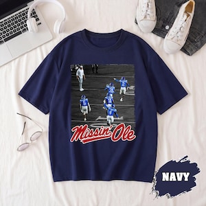 May include: A navy blue short-sleeve t-shirt featuring a graphic print of football players in blue uniforms on a field. The text "Missin' Ole" is printed in red script below the players. The shirt is displayed on a white surface.