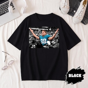 May include: A black short-sleeved t-shirt featuring a central graphic print of a football player wearing a teal jersey with the number 39, surrounded by teammates in a stadium setting, with a white signature above the player's head.