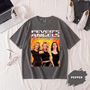May include: A charcoal gray short-sleeve t-shirt featuring a graphic print of three individuals in black tops against a fiery orange background. The text FEVER'S ANGELS FULL THROTTLE is printed in white and silver above the figures.