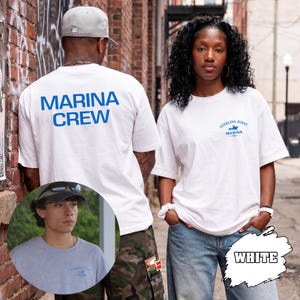 May include: A white short-sleeved t-shirt featuring the blue text MARINA CREW on the back and a small blue graphic with the text STERLING POINT MARINA on the front left chest. The shirt is shown in a composite image with a grey version of the same shirt.