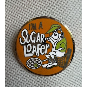 Puede incluir: Un pin esmaltado redondo con fondo naranja que muestra un personaje de dibujos animados con visera y camiseta verdes, sosteniendo una raqueta de tenis con una pelota. El texto I'M A SUGAR LOAFER aparece en letras blancas y negras.