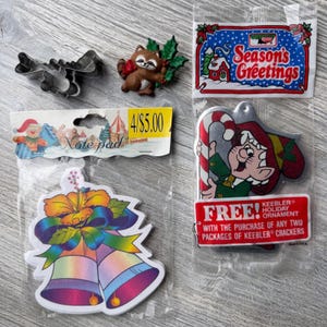 May include: A metal cookie cutter, a raccoon pin with green leaves, a note pad with a colorful bell illustration, and a Keebler holiday ornament in plastic packaging. The ornament features an elf with a candy cane and text reading FREE! KEEBLER HOLIDAY ORNAMENT.
