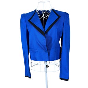 May include: A vibrant royal blue cropped blazer featuring a sharp notched collar with black trim, long sleeves with black cuffs, and a single front button closure. The jacket has a structured shoulder design and two horizontal welt pockets at the waist.