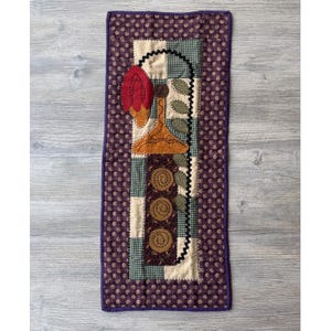May include: A rectangular quilted wall hanging featuring a dark purple fabric with a small floral pattern. The center panel displays a patchwork design with cream, green checkered, and brown fabrics, decorated with appliqued shapes of a red flower, a gold vase, green leaves, and three brown circular motifs.