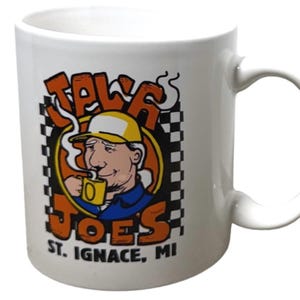 May include: A white ceramic mug featuring a graphic of a person in a yellow cap drinking from a yellow mug. The text reads JALA JOES ST. IGNACE, MI in orange and black lettering, set against a black and white checkered border on a circular background.