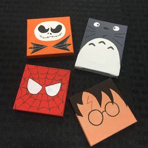 Puede incluir: Cuatro lienzos cuadrados pintados con personajes de dibujos animados. Incluye a Jack Skellington sobre naranja, Totoro sobre gris, la máscara de Spider-Man sobre rojo y Harry Potter con gafas y cicatriz.