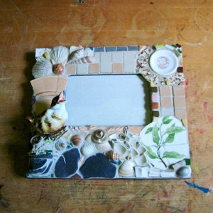 May include: A mosaic picture frame with a bird figurine, seashells, and a small plate with a floral design. The frame has a light blue background and a white mat.