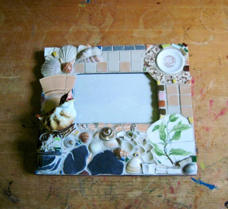 May include: A mosaic picture frame with a bird figurine, seashells, and a small plate with a floral design. The frame has a light blue background and a white mat.