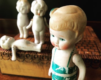 May include: Four porcelain dolls, three standing and one lying down. The dolls are all white with painted features and hair. One doll is wearing a green and white swimsuit.