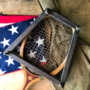 May include: A vintage wooden tennis racket with a brown leather grip and a stringed frame. The racket is partially obscured by an American flag and a brown canvas bag.