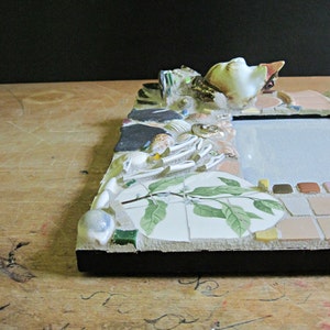 May include: A picture frame made of mosaic tiles with a white porcelain bird figurine on top. The frame is decorated with seashells, leaves, and other natural elements. The frame is set on a wooden surface.