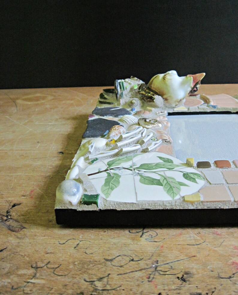 May include: A picture frame made of mosaic tiles with a white porcelain bird figurine on top. The frame is decorated with seashells, leaves, and other natural elements. The frame is set on a wooden surface.