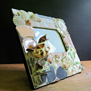 May include: A mosaic picture frame with a bird figurine, seashells, and other decorative elements. The frame is made of various materials, including ceramic tiles, glass, and metal.