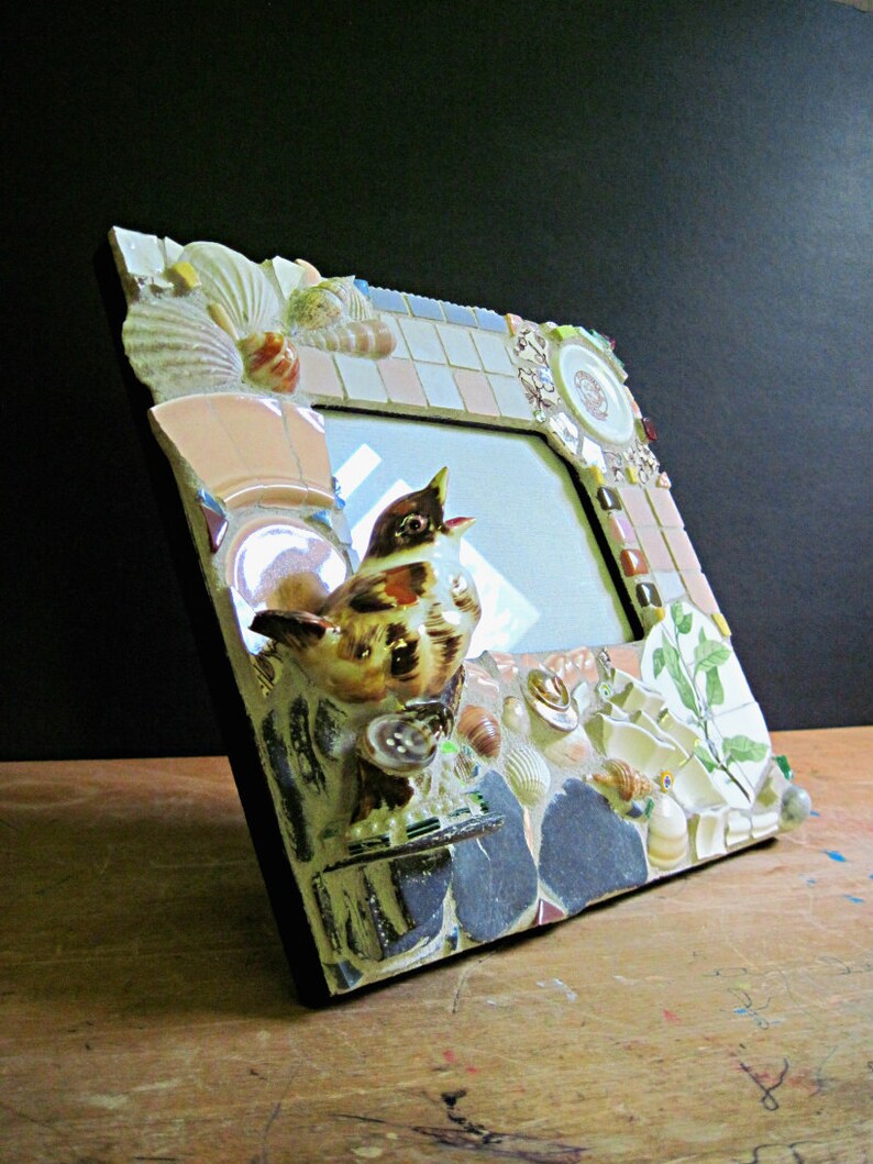 May include: A mosaic picture frame with a bird figurine, seashells, and other decorative elements. The frame is made of various materials, including ceramic tiles, glass, and metal.