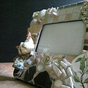 May include: A white ceramic bird figurine sits on a mosaic picture frame. The frame is made of various materials, including shells, tiles, and glass. The frame has a white mat and a clear glass front.