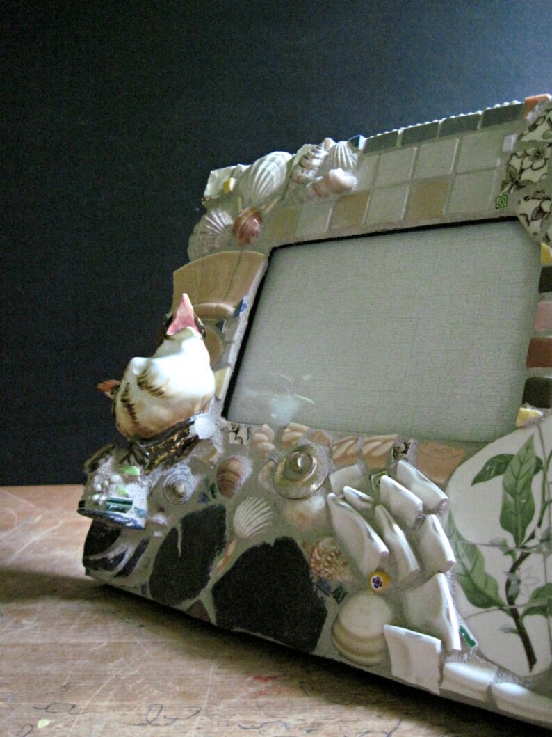 May include: A white ceramic bird figurine sits on a mosaic picture frame. The frame is made of various materials, including shells, tiles, and glass. The frame has a white mat and a clear glass front.
