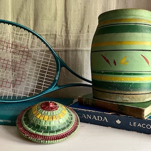May include: A green and yellow striped ceramic jar with red and yellow painted leaves. The jar is sitting on top of a stack of books with the word "CANADA" printed on the top book. A blue tennis racket and a green, yellow, and red circular object are also in the image.