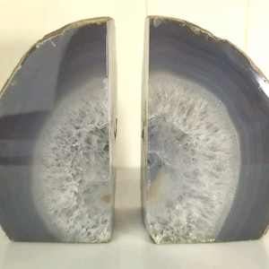 Peut inclure: Une paire de serre-livres fabriqués à partir de géodes d'agate tranchées. Chaque pièce présente une face polie, lisse et semi-circulaire affichant des bandes concentriques de tons bleu et gris sourds entourant un amas central de cristaux blancs translucides.