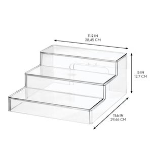 May include: A clear, three-tiered acrylic display stand with a stepped design. The unit measures 11.2 inches in width, 11.6 inches in depth, and 5 inches in height, providing three distinct levels for organizing items in a tiered arrangement.