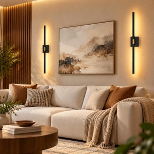 May include: Two vertical wall sconces featuring a slim black rod design with a central gold accent, mounted on either side of a landscape painting. The lights emit a warm glow against the wall, illuminating the space above a beige sofa.