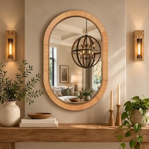 May include: An oval wall mirror with a light brown, textured wooden frame. The mirror reflects a room interior featuring a spherical metal chandelier, a white sofa with cushions, a table lamp, and a framed picture on the wall behind the mirror.