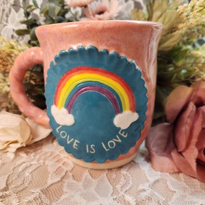 Puede incluir: Una taza de cerámica rosa con un diseño de arcoíris azul y blanco. La taza tiene las palabras "Love is Love" escritas en ella.