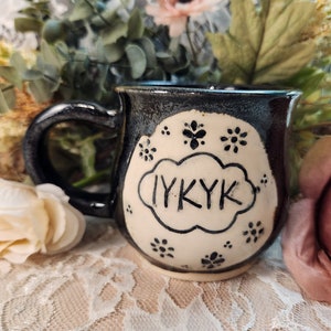 Puede incluir: Una taza de cerámica negra y crema con asa. La taza presenta el texto "IYKYK" en un diseño en forma de nube, rodeado de pequeños detalles florales. La taza está sobre un fondo floral.