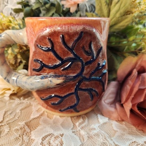 May include: A handmade ceramic mug with a unique design. The mug features a textured, twisted handle and a reddish-brown body with a dark blue, branch-like pattern. The rim has an orange hue.