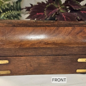 May include: A rectangular wooden chest with a curved lid, featuring polished brass corner accents and brass plates on the front. The wood displays a natural grain pattern in shades of brown. The word FRONT is printed in a white box at the bottom.