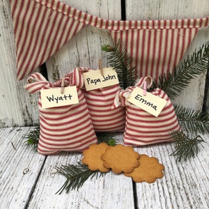 May include: Three red and white striped fabric gift bags with the names Wyatt, Papa John, and Emma written on them. The bags are sitting on a white wooden surface with three gingerbread cookies.