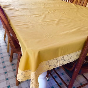 May include: A yellow tablecloth with a lace trim. The tablecloth is on a wooden table with white legs. The table is in a room with a tiled floor.