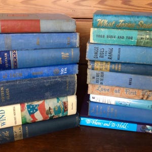May include: A collection of vintage books in various shades of blue, stacked on a wooden surface. Titles include "Gone with the Wind", "Ride with Me", and "What Jesus Said". The books have aged covers and spines, showcasing their vintage appeal.