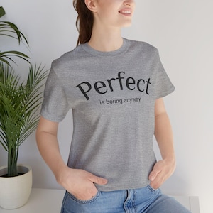 Op de afbeelding: Een grijs gemêleerd T-shirt met korte mouwen en de zwarte tekst "Perfect is boring anyway" op de borst gedrukt. Het shirt heeft een ronde hals en een standaard pasvorm, gemaakt van een zachte stof met een zichtbare gemêleerde textuur.
