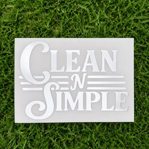 Może przedstawiać: Prostokątna biała naklejka z napisem Clean N Simple wykonanym pogrubioną, stylizowaną białą czcionką. Słowo Clean znajduje się nad słowem Simple, z mniejszą literą N pośrodku, otoczoną z obu stron trzema poziomymi liniami.
