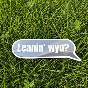 Può includere: Un adesivo a forma di fumetto con uno sfondo sfumato grigio scuro e un bordo bianco. Il testo "Leanin' wyd?" è stampato in grassetto bianco senza grazie al centro dell'adesivo, imitando l'aspetto di un messaggio digitale.