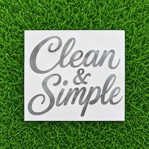 May include: A square white decal featuring the dark gray text Clean & Simple in a cursive script font. The lettering is centered on the white background, with the word Clean on the top line and the word Simple on the bottom line.