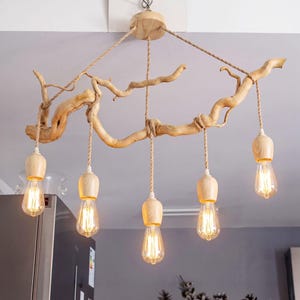 Pode incluir: Uma luminária de teto com um galho de madeira de cor clara natural suspenso por uma corda grossa. Cinco soquetes com coberturas de madeira correspondentes pendem do galho, cada um equipado com uma lâmpada brilhante estilo Edison.